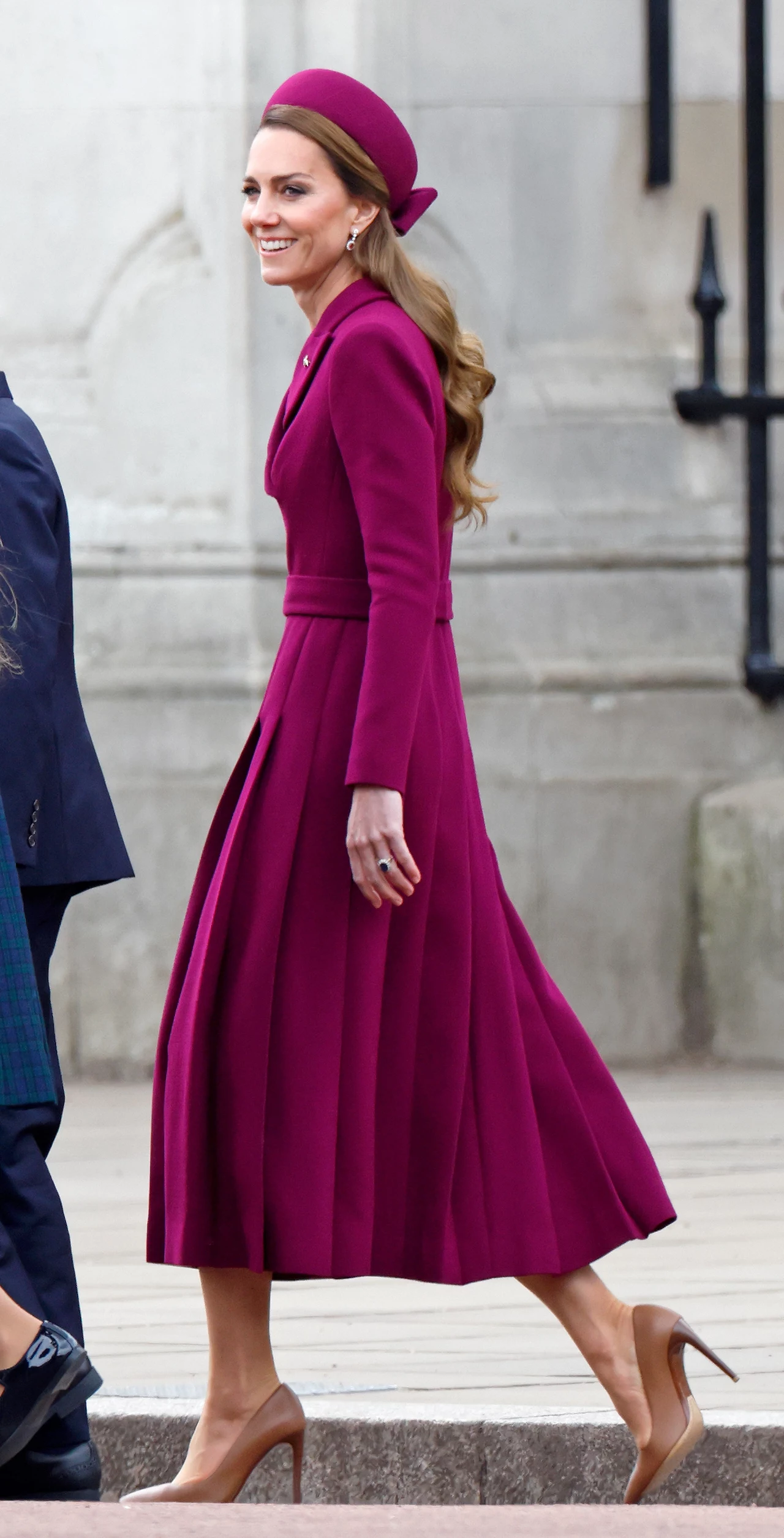 Magenta to kolor dla osób, które lubią być w centrum uwagi Elegancko ubrana kobieta w długiej, bordowej sukience oraz kapeluszu w tym samym kolorze, idąca po chodniku. Obok niej widoczne są inne osoby, niewidoczne w całości.