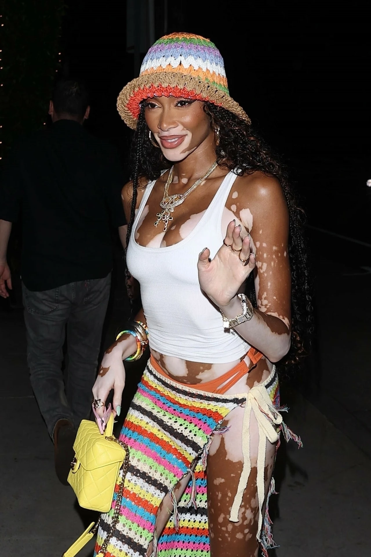 Modelka Winnie Harlow w kolorowym kapeluszu rybaka Uśmiechnięta kobieta z ciemnymi, kręconymi włosami w kolorowym kapeluszu i białym topie. Na szyi ma naszyjnik, w ręku żółtą torebkę. Część garderoby to kolorowa spódniczka. Na skórze widoczne jasne plamy, schorzenie znane jako bielactwo.