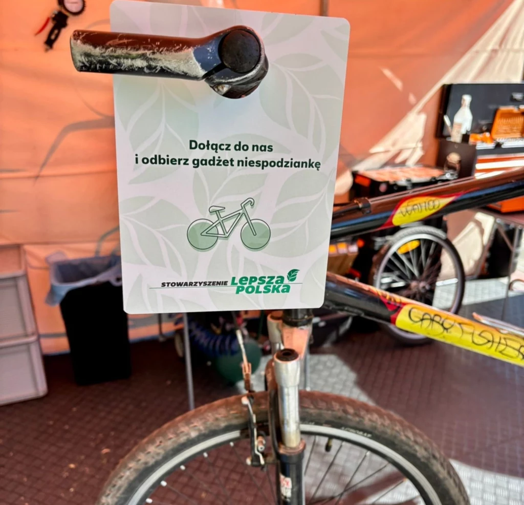Akcja "Zmień 4 kółka na rower" Rower z zamontowaną kartką zachęcającą do dołączenia do stowarzyszenia 'Lepsza Polska' w zamian za gadżet niespodziankę. W tle namiot, w którym widać stojące przedmioty związane z akcją promocyjną.