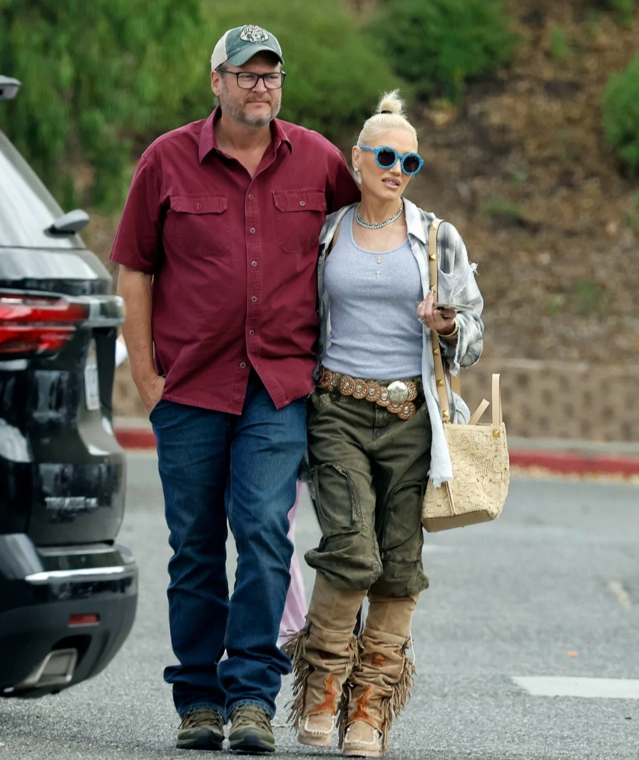 Gwen Stefani z mężem, Blakiem Sheltonem Mężczyzna w czerwonej koszuli i czapce oraz kobieta w okularach przeciwsłonecznych i luźnej stylizacji spacerują razem. Kobieta trzyma koszyk, a para wydaje się być w przyjaznej relacji.