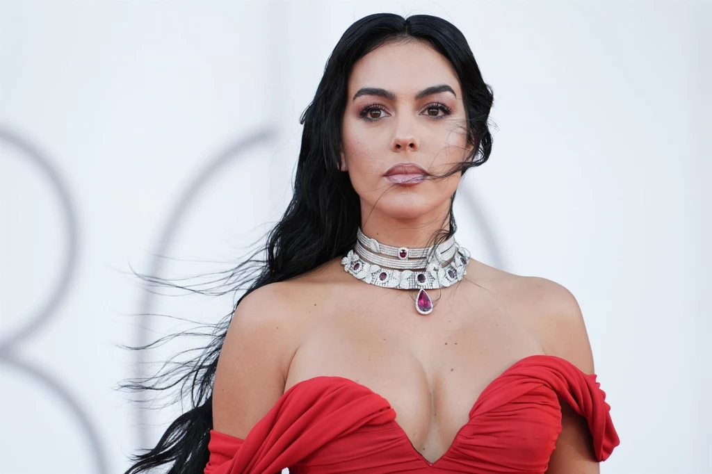 Georgina Rodriguez kocha bogatą biżuterię Kobieta ubrana w czerwoną suknię z odkrytymi ramionami, ozdobiona wyrazistą biżuterią z dużym kamieniem, włosy opadają swobodnie, na jasnym tle.