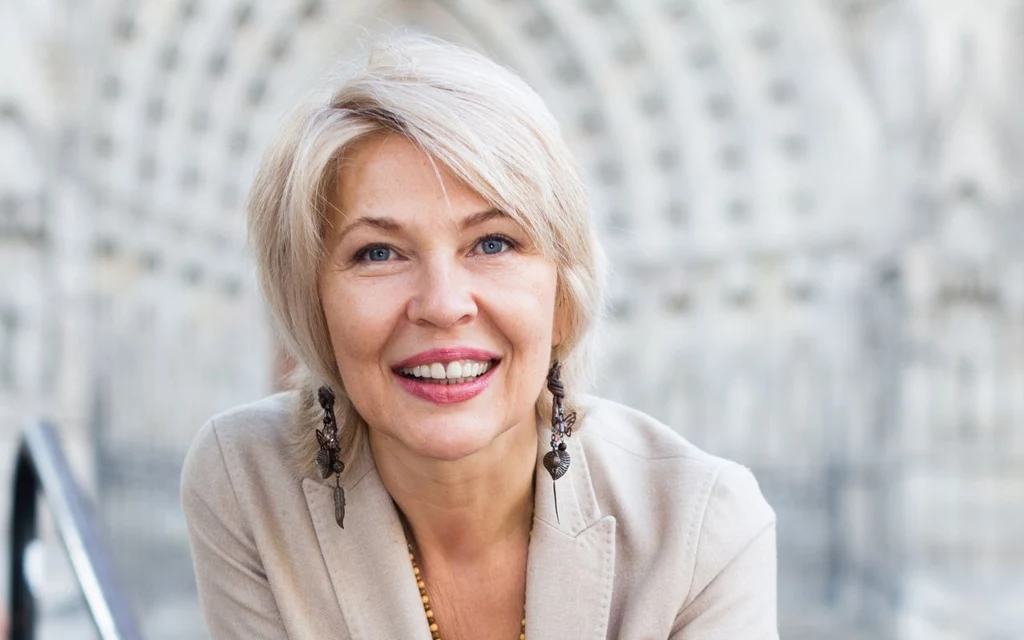 Jak dobrać fryzurę do wieku? Cieniowanie może nadać twarzy świeżości Uśmiechnięta kobieta z krótkimi blond włosami w beżowej marynarce, stojąca na zewnątrz. Tło rozmyte, architektoniczne detale. Kobieta ma na sobie długie kolczyki.