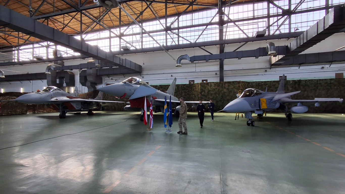 Samoloty myśliwskie w dużym hangarze, pośrodku widoczni są ludzie w mundurach. Jasne światło wpadające przez duże okna na suficie oświetla wnętrze.