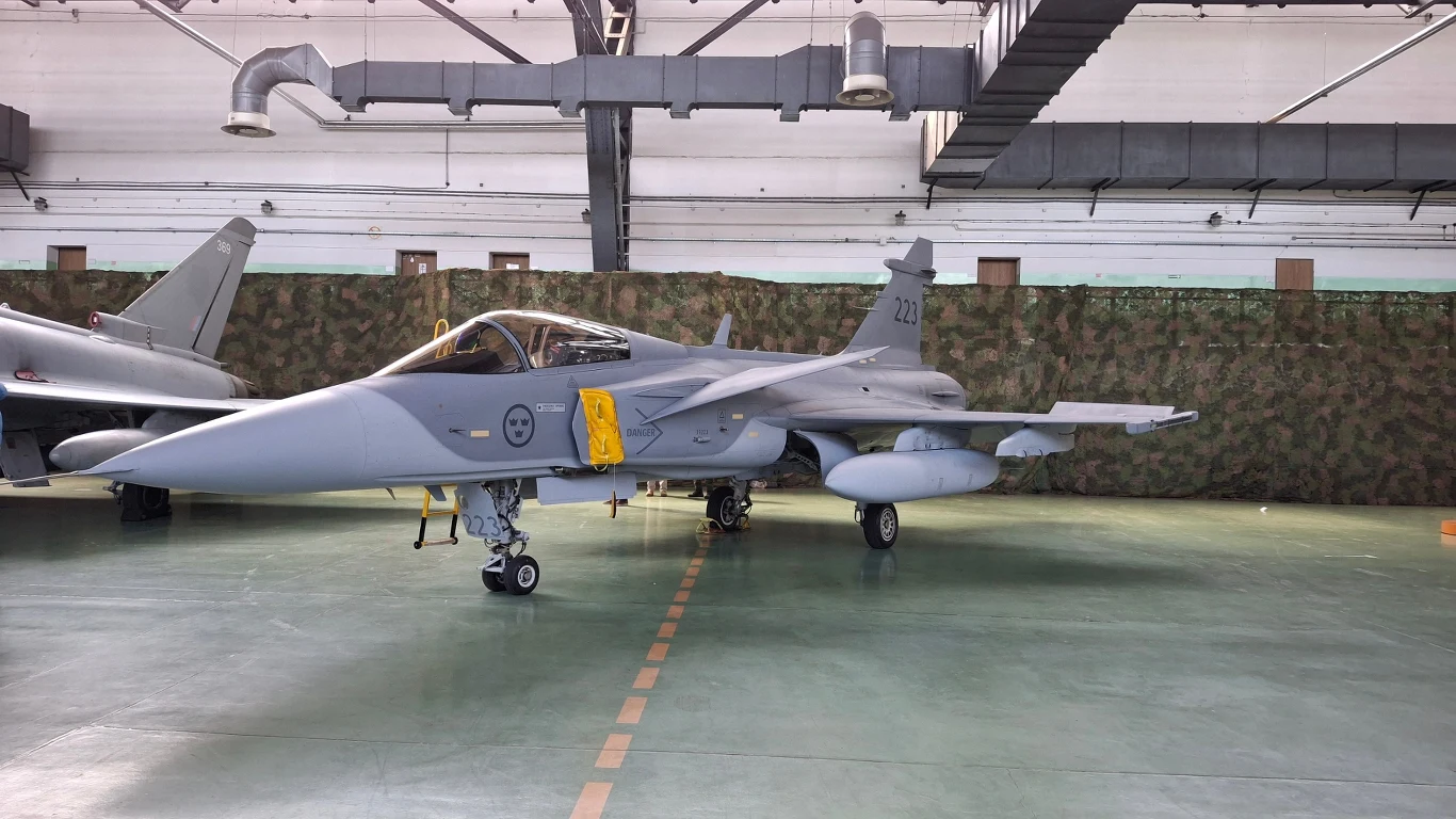 Myśliwiec stojący w hangarze, otoczony delikatnym światłem. Widoczny jest szczegółowy przód samolotu z oznaczeniami i wyposażeniem.