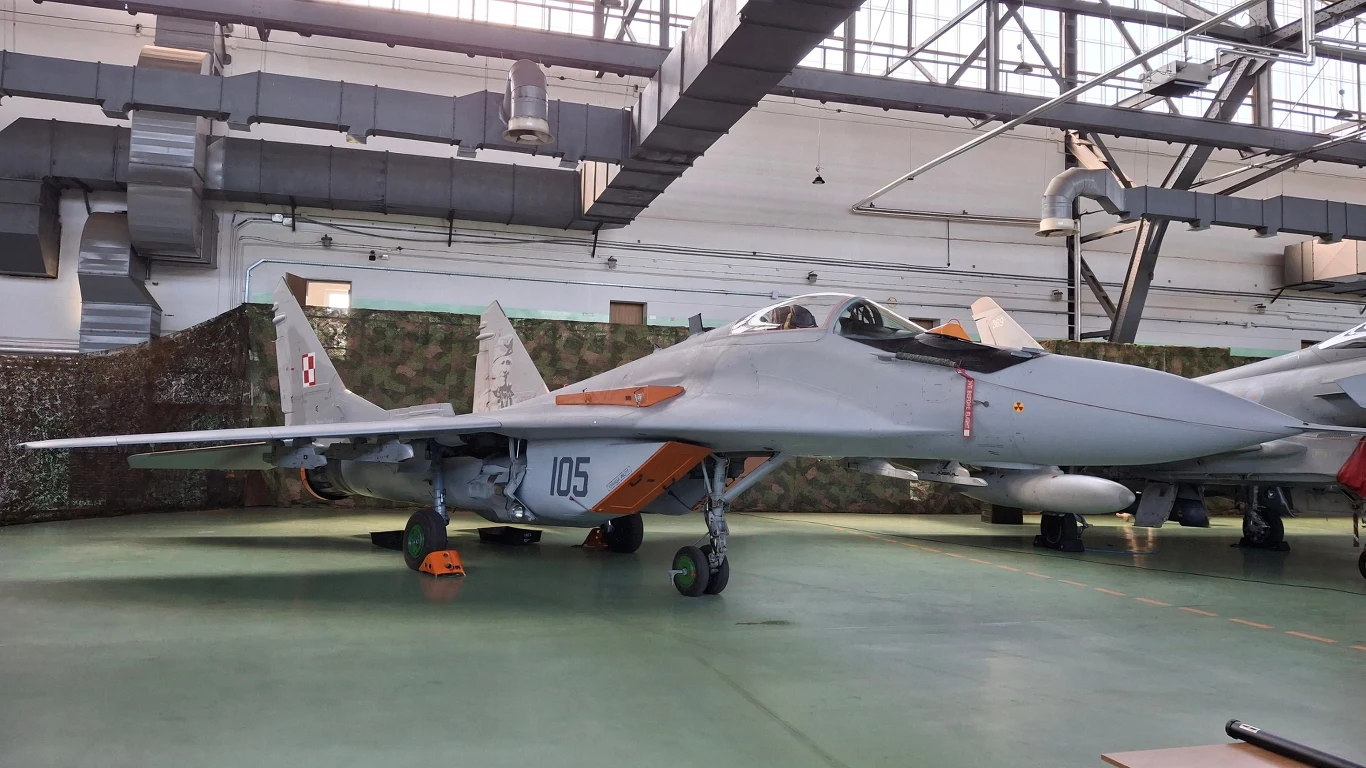 Samolot myśliwski MiG-29 w hangarze, oznakowany numerem 105. Maszyna stoi w przestronnym, dobrze oświetlonym budynku z dużymi stalowymi konstrukcjami sufitowymi.