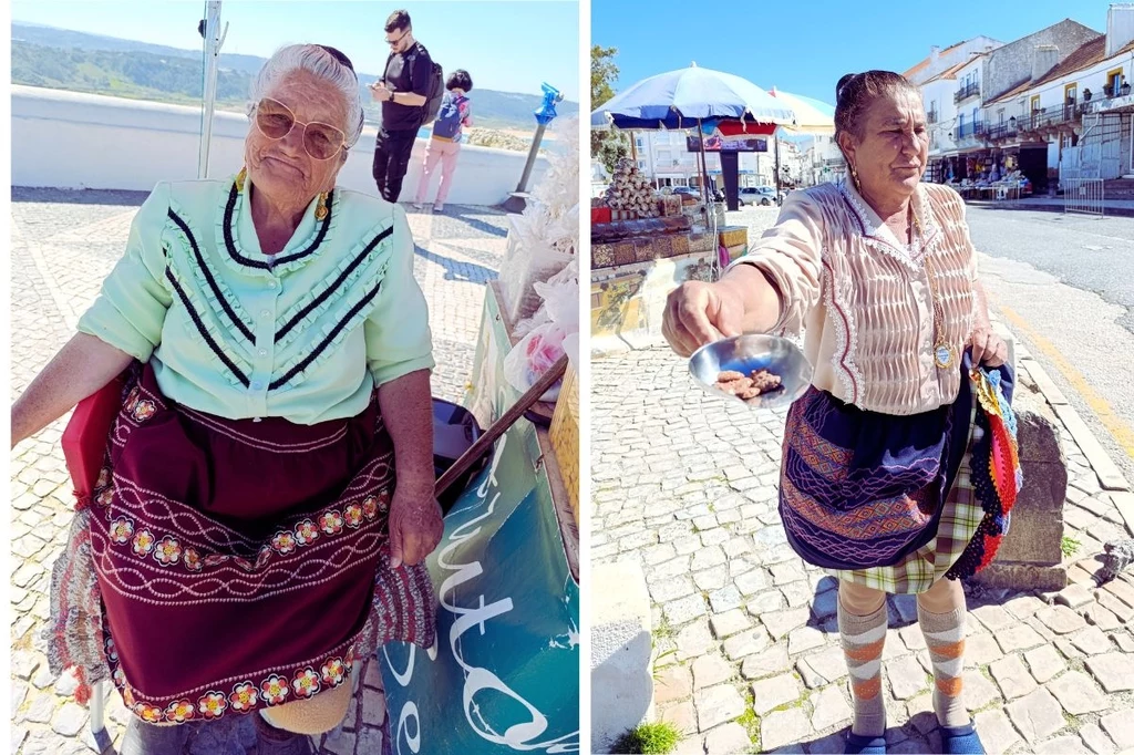 Mieszkanki Nazaré w tradycyjnych strojach Starsze kobiety w tradycyjnych, kolorowych strojach pozują na ulicy, jedna siedzi, druga stoi z wyciągniętą ręką. W tle widać jasne budynki i niebieskie niebo.