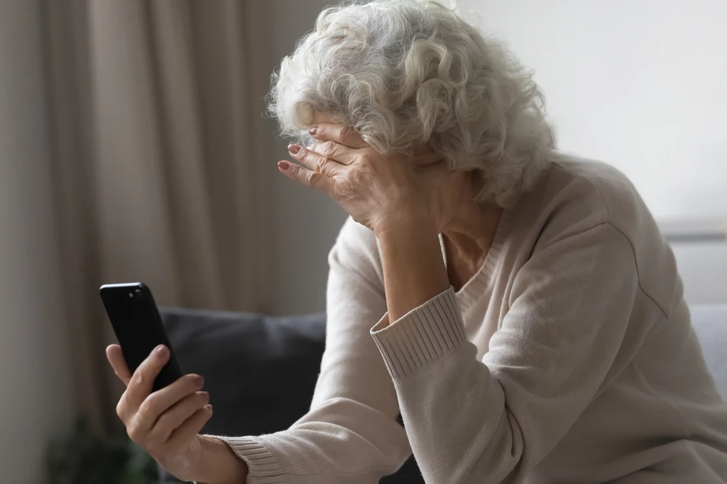 Jeśli oszuści nie uzyskają dostępów przez internet, zaczynają dzwonić do swoich ofiar Siedząca kobieta w średnim wieku o siwych włosach trzyma telefon komórkowy w dłoni i zasłania twarz drugą ręką. Jest ubrana w jasny sweter, siedzi na kanapie w domowym wnętrzu. Wygląda, jakby była zmartwiona lub przygnębiona.