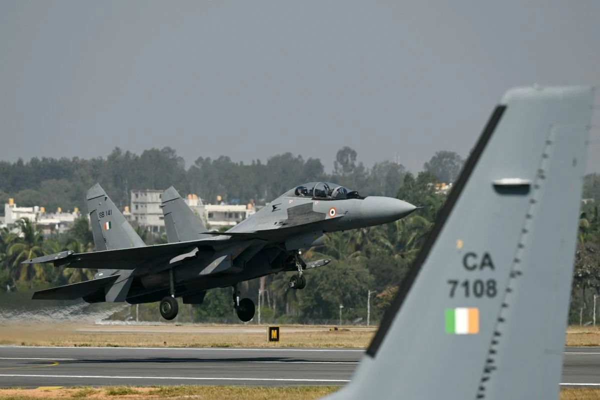 Samolot wielozadaniowy Su-30MKI indyjskich sił powietrznych Samolot wojskowy w trakcie lądowania na pasie startowym. W tle znajdują się drzewa oraz budynki, a na pierwszym planie widoczny jest fragment ogona innego samolotu z oznaczeniem i flagą Indii.