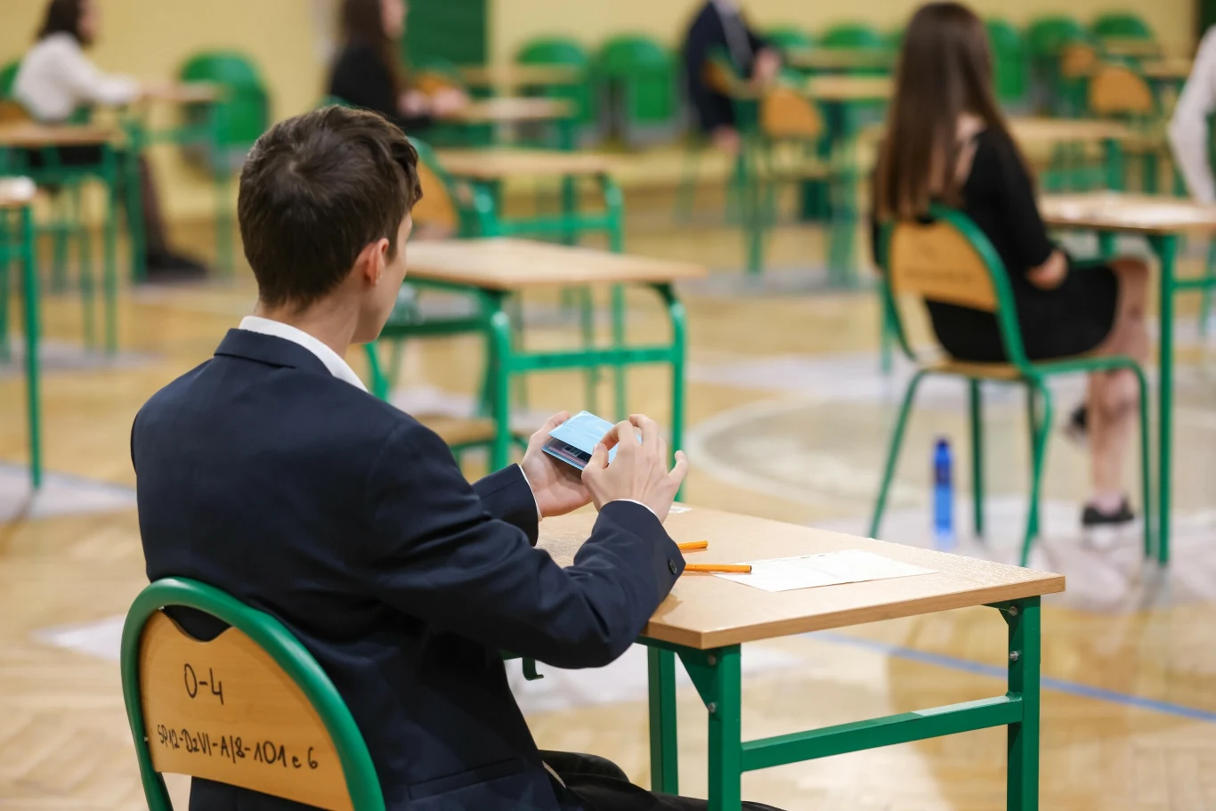 Egzaminy ósmoklasistów - jak się odwołać od wyniku? Uczeń przy ławce podczas egzaminu ósmoklasisty, w tle inni uczniowie w sali egzaminacyjnej.
