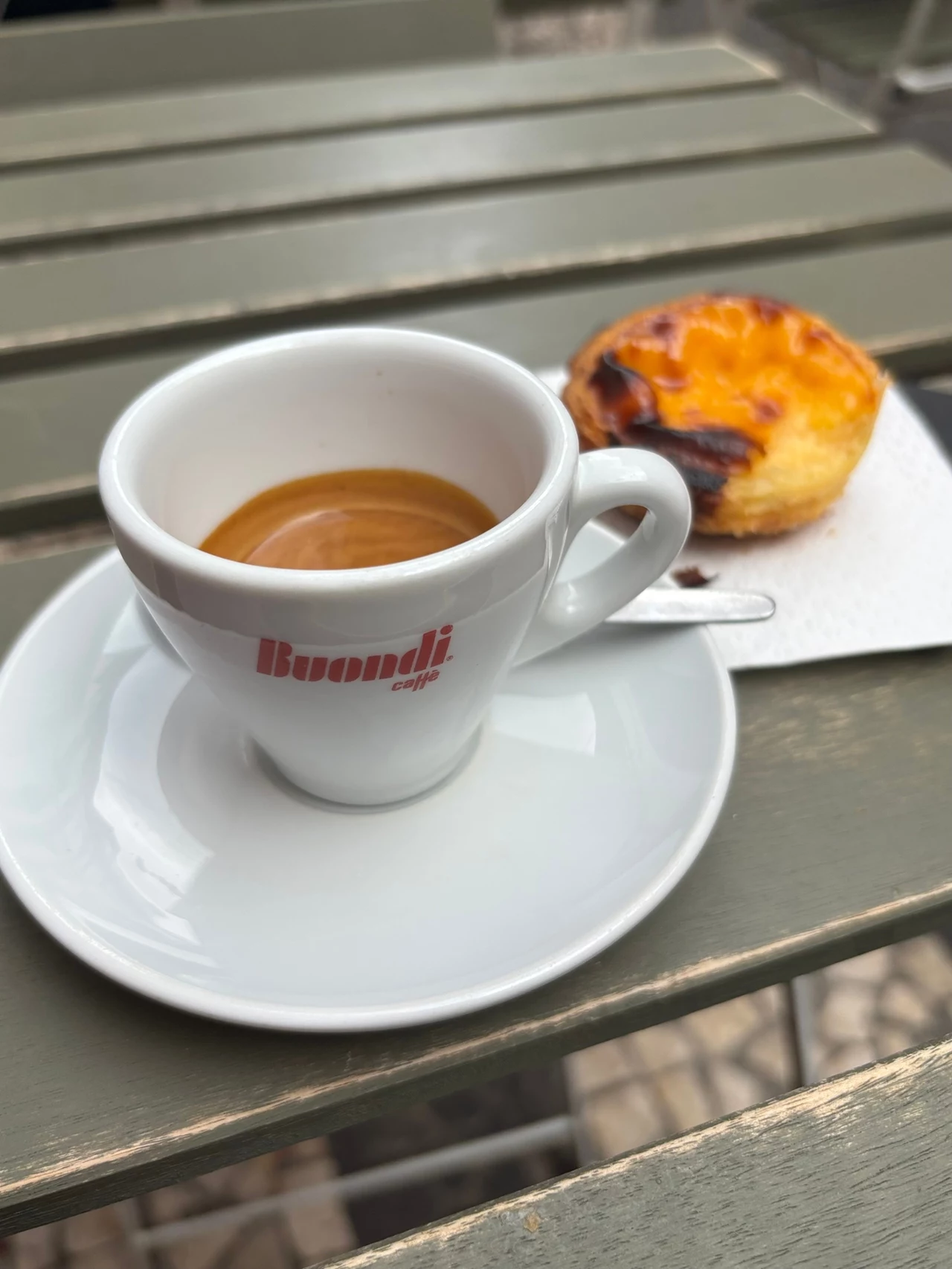Kawa i ciasteczko to koszt ok. 2 euro. Filiżanka espresso Buondi na metalowym stoliku wraz z pastel de nata. Kubek jest biały z czerwonym logo. Ciastko znajduje się na białej serwetce, lekko przypieczone, z kremowym wnętrzem.