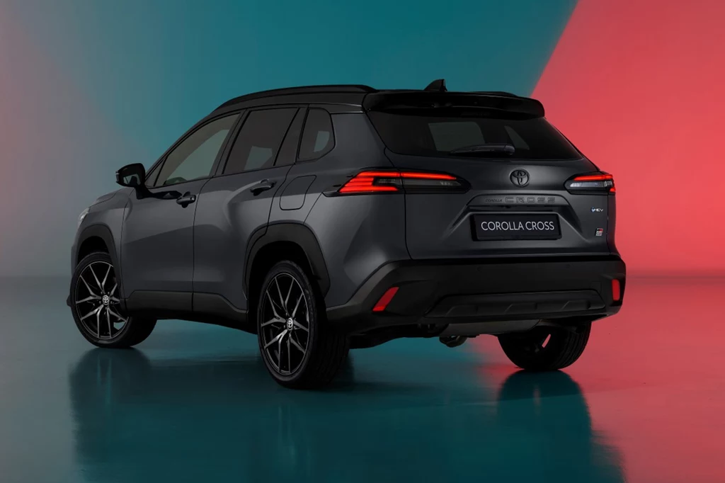 Z tyłu zauważymy subtelną zmianę – logo Corolli Cross zostało wtłoczone, a lampy zyskały nowy design. Ciemny samochód SUV Toyota Corolla Cross na kolorowym tle, widziany z tyłu i z boku, podkreślający elegancki design pojazdu. Tło w odcieniach niebieskiego i czerwonego tworzy kontrast, uwydatniając sylwetkę auta.
