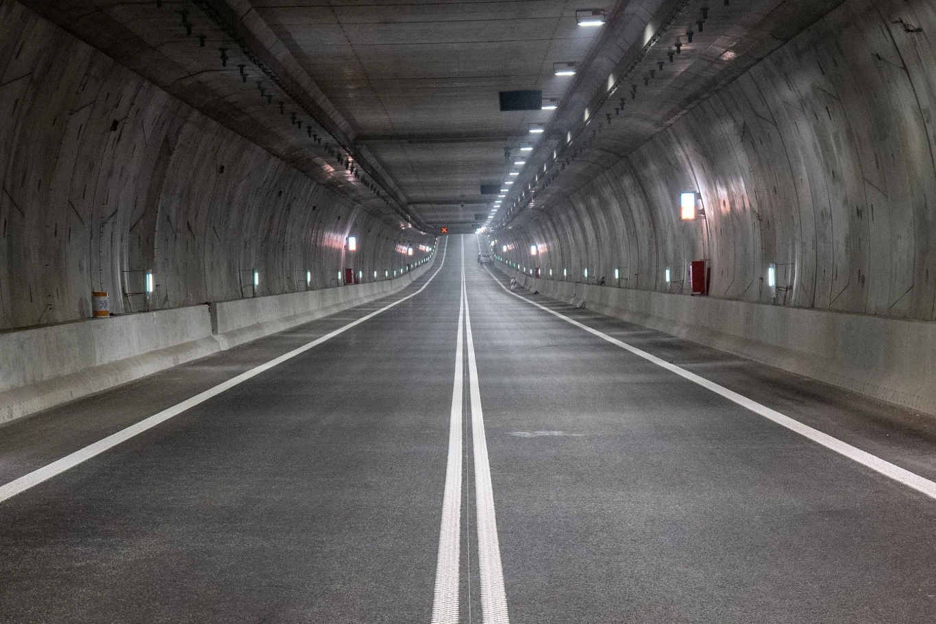 Pusty, dobrze oświetlony tunel drogowy z wyraźnie zaznaczonymi liniami na asfalcie. Ściany tunelu są betonowe, oświetlenie równomiernie rozłożone na całej długości.