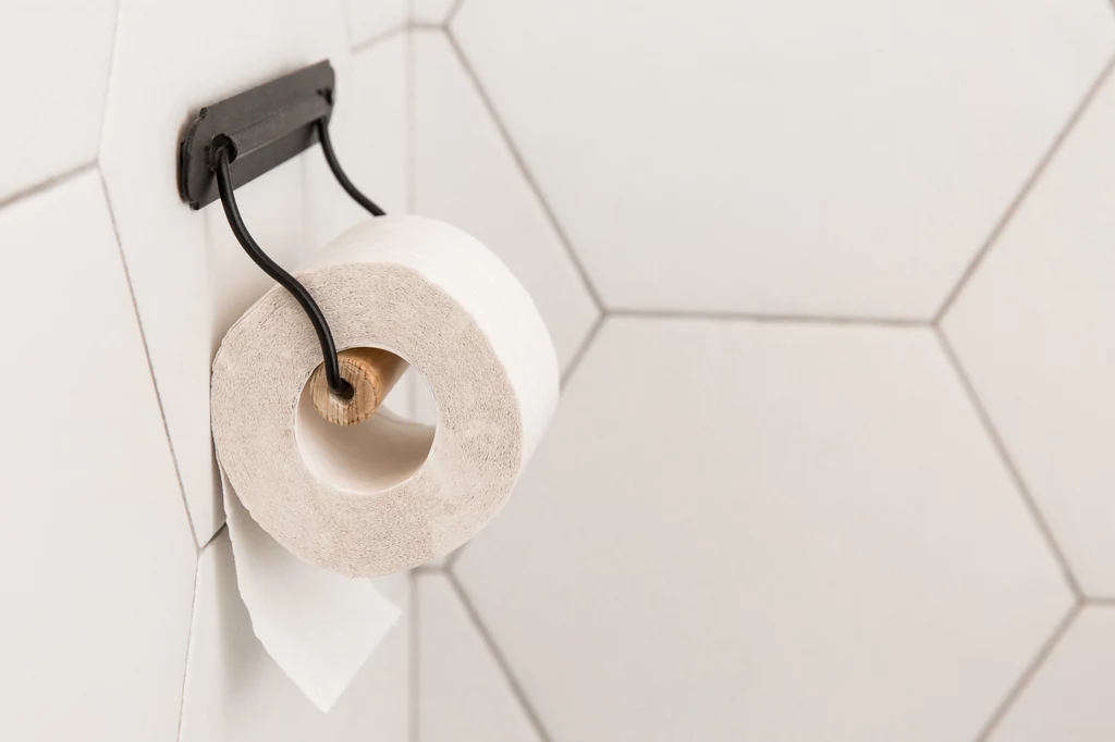 Zaledwie 30% osób trzyma papier toaletowy rozwijający się od tyłu. Rolka papieru toaletowego zawieszona na czarnym uchwycie na tle białych, hexagonalnych płytek ściennych.