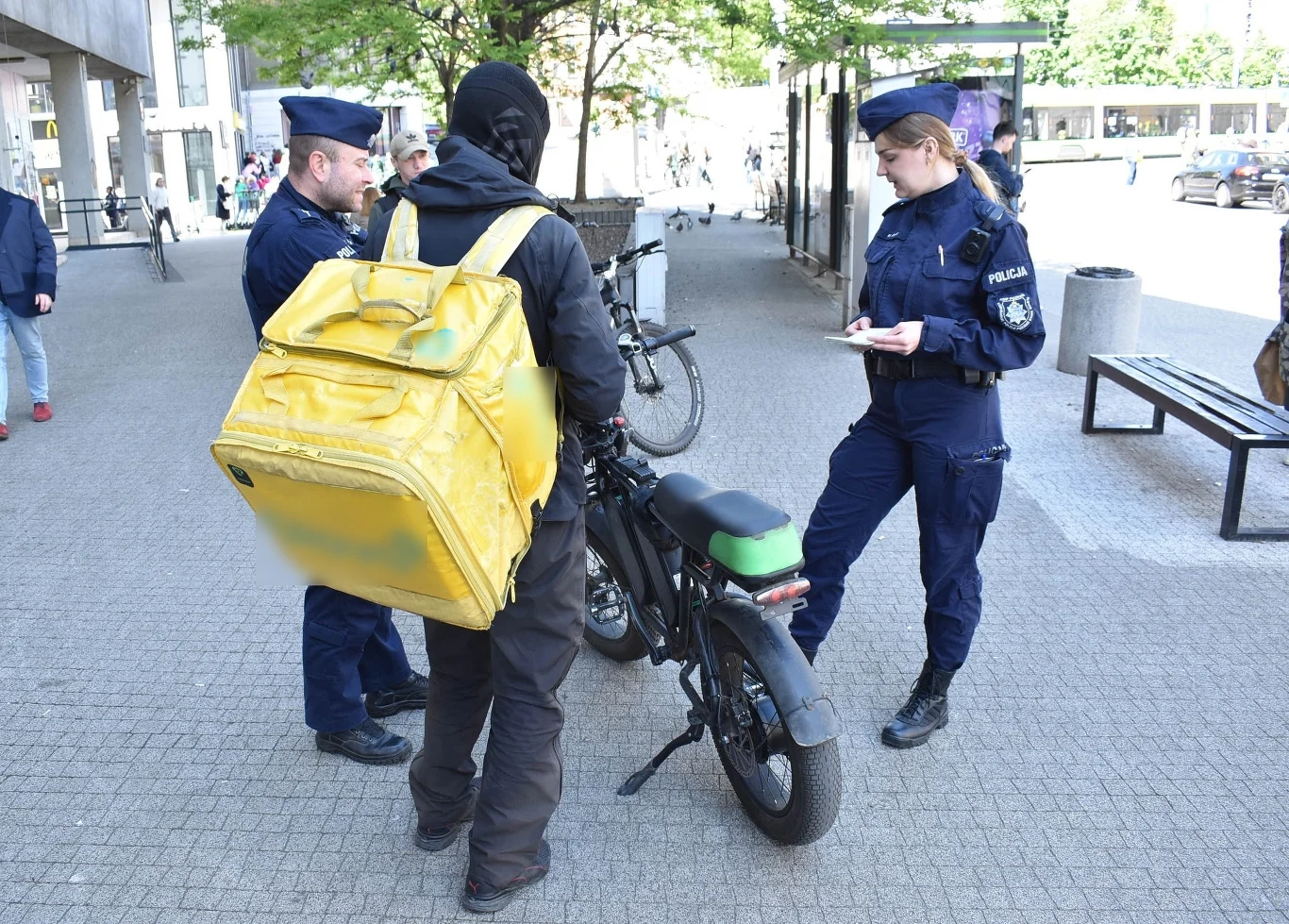 Rower elektryczny musi spełniać definicję określoną w przepisach Policjanci zatrzymują mężczyznę z żółtym plecakiem i rowerem elektrycznym na chodniku miejskim. Funkcjonariuszka sporządza notatki, podczas gdy dwóch innych policjantów obserwuje sytuację. W tle widoczne są budynki i przechodnie.