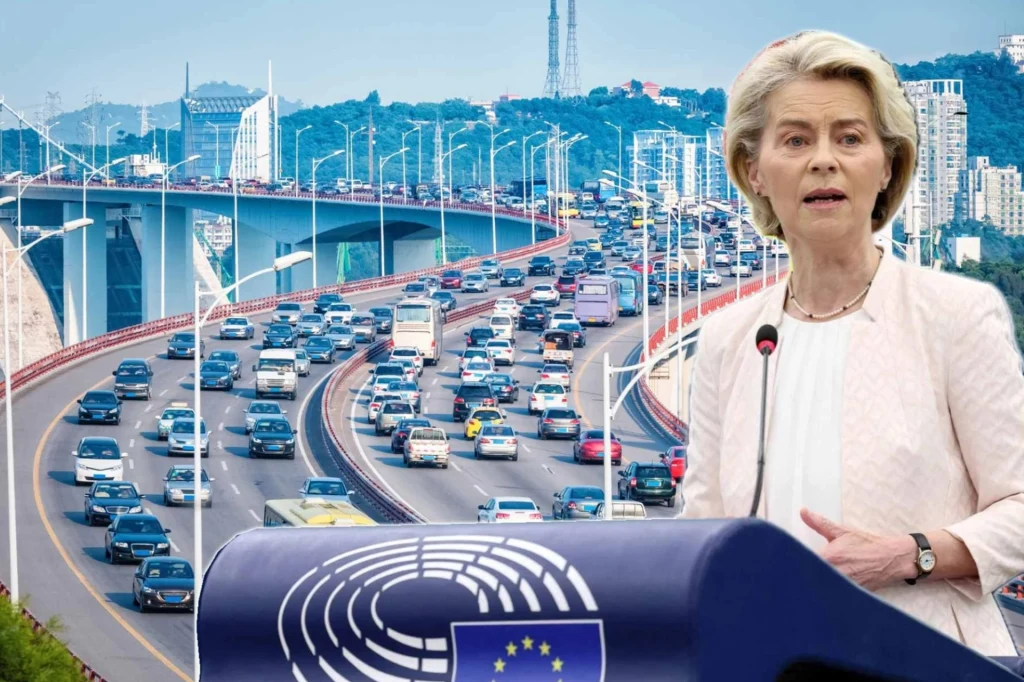 Osoba przy mównicy z logo Unii Europejskiej na tle ruchliwego mostu z dużym natężeniem ruchu samochodowego. Mównicę zdobi emblematu unijny, co sugeruje kontekst oficjalnego wystąpienia.