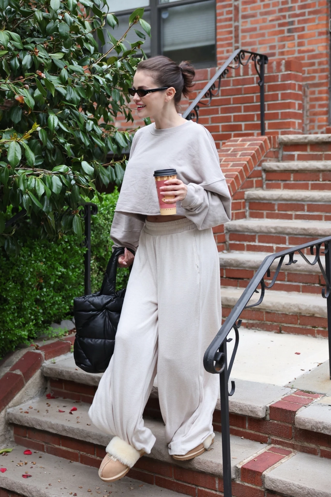 Selena Gomez w wygodnej stylizacji Kobieta w jasnym stroju w stylu casual idzie po schodach z kubkiem kawy w ręku. Ma na sobie ciemne okulary przeciwsłoneczne, a w drugiej ręce trzyma czarną torbę. Tło stanowi ceglana ściana oraz zielony krzew.