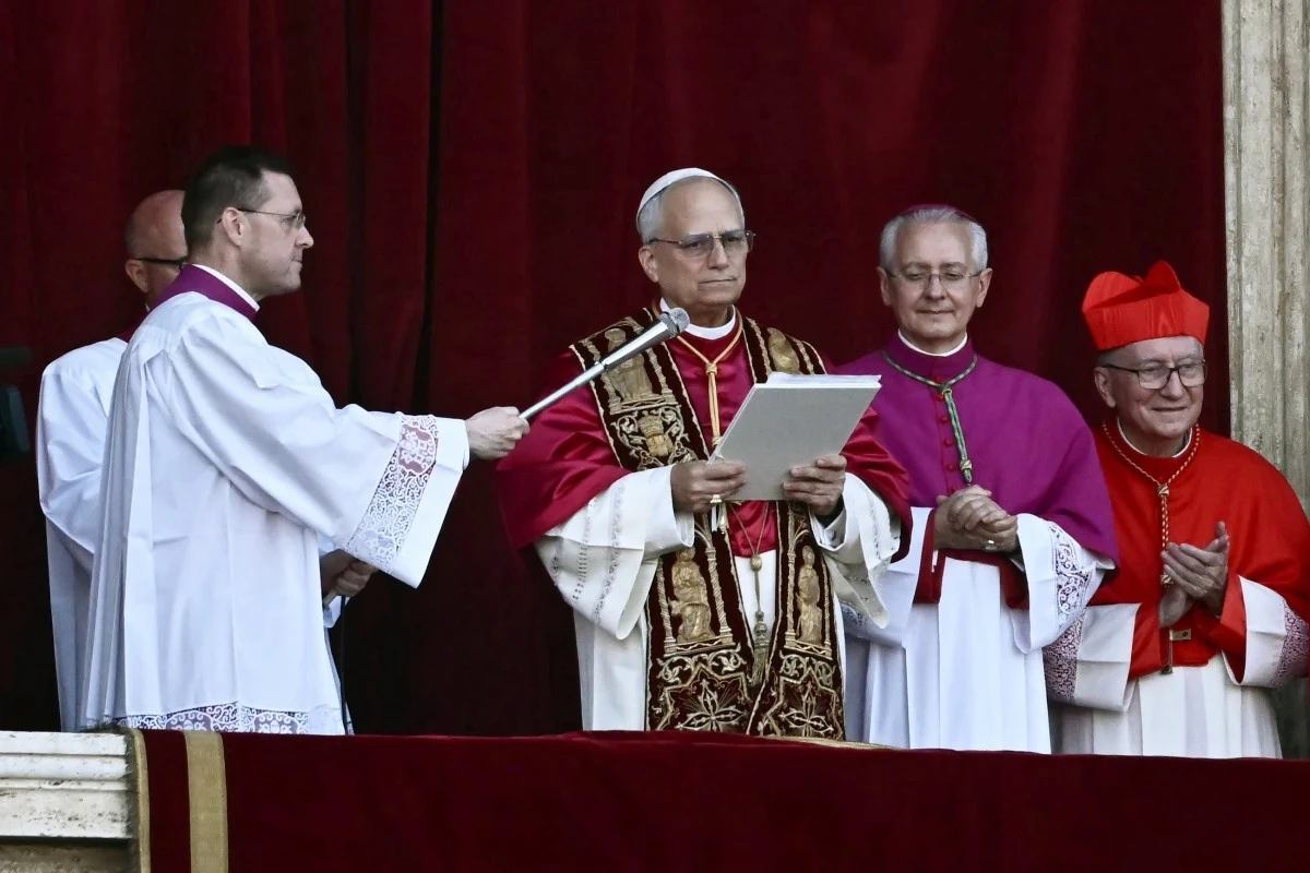 Duchowni w strojach liturgicznych na balkonie, jeden z nich trzyma dokument, a drugi mikrofon. Tło stanowi czerwona zasłona.