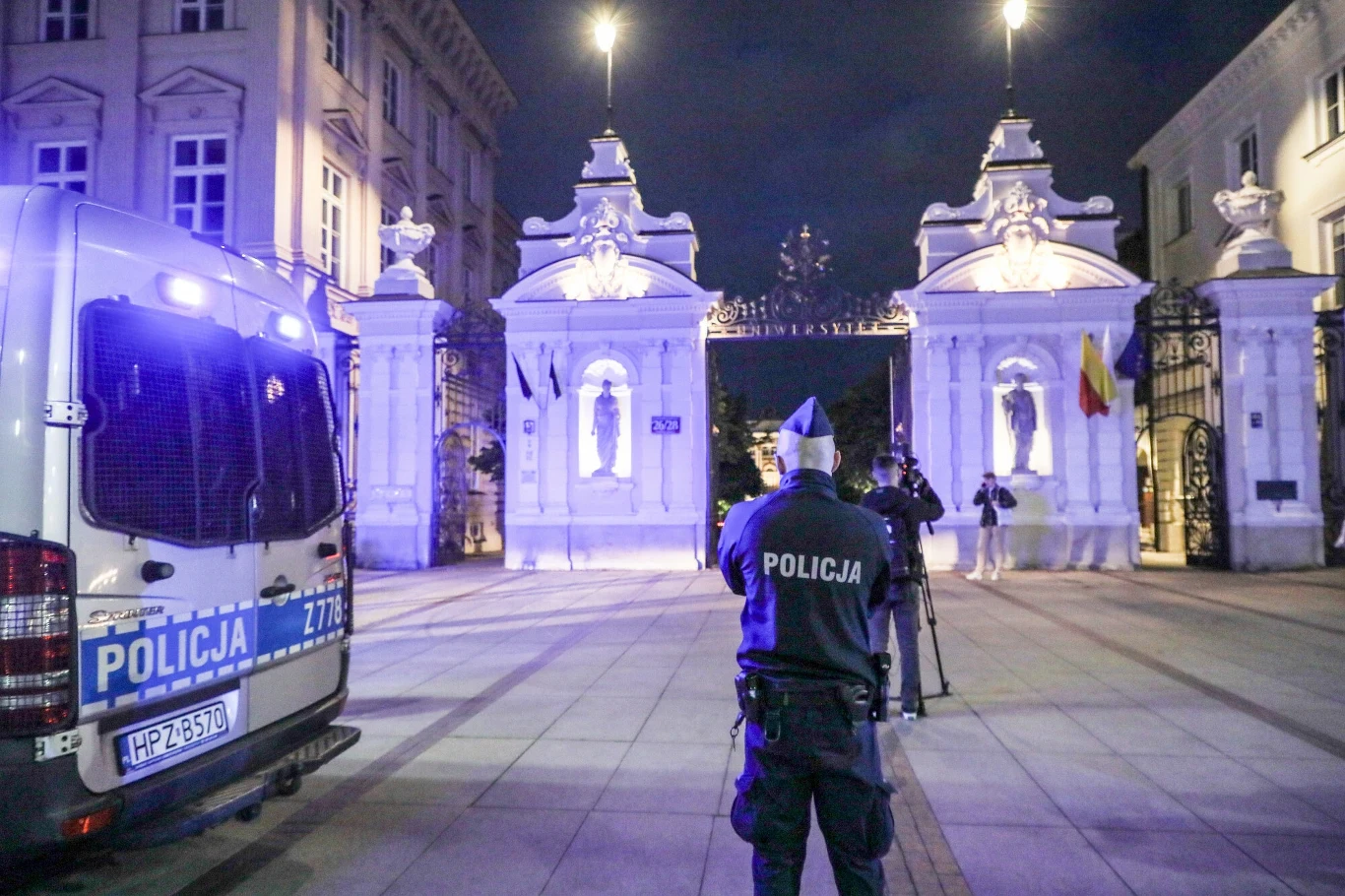 Zabójstwo na kampusie UW. Student przyznał się do winy Policjant patroluje obszar przed główną bramą Uniwersytetu Warszawskiego nocą. W tle widoczna jest ozdobna brama i policyjny pojazd z włączonymi światłami.