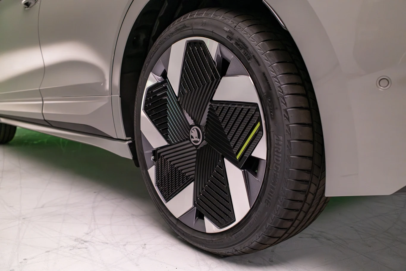 Koło samochodowe z nowoczesnym felgiem o geometrycznym wzorze, widoczne na tle podłogi w neutralnym kolorze, bliskość samochodu sugeruje nowe technologie i design.