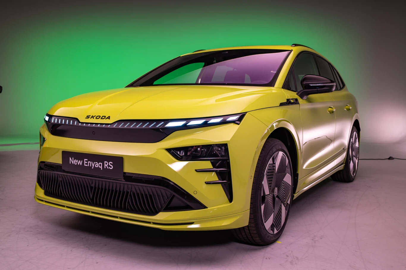 Jasnozielony samochód Škoda Enyaq RS na neutralnym tle, z zielonym oświetleniem w tle. Samochód ma nowoczesny, sportowy design i charakterystyczne przednie reflektory LED. Widoczna tabliczka z napisem 'New Enyaq RS'.