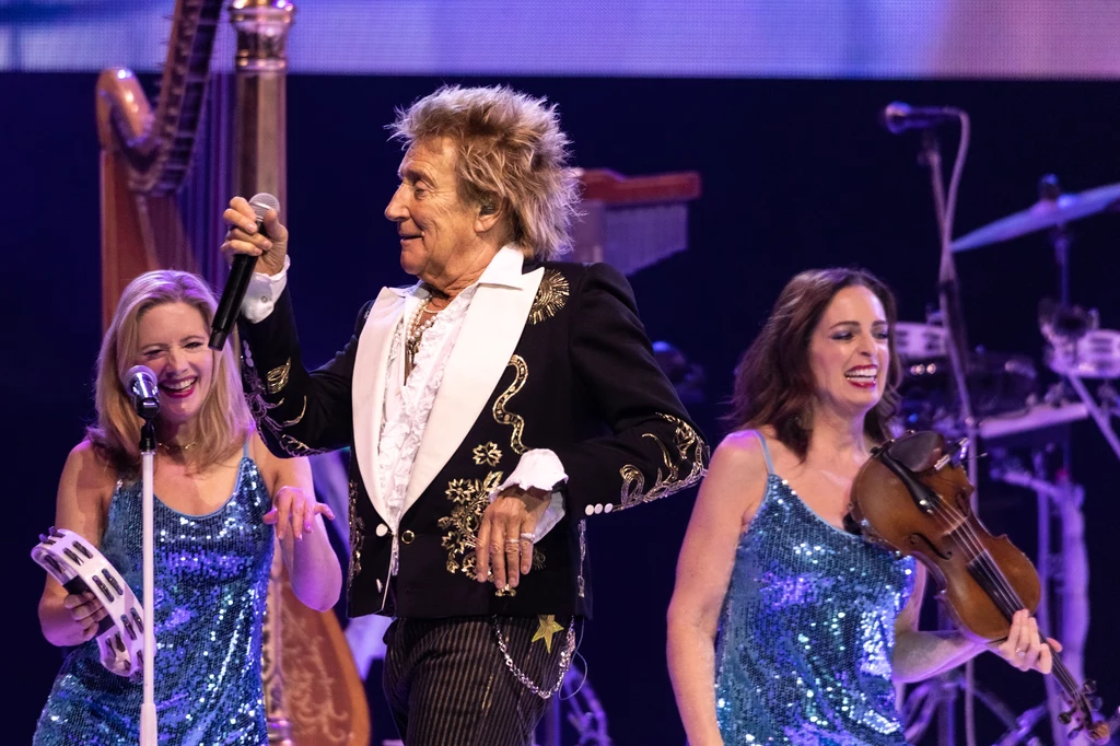 Rod Stewart w Tauron Arenie Kraków Mężczyzna w eleganckim stroju śpiewa na scenie, trzymając mikrofon. Obok niego dwie kobiety grające na instrumentach muzycznych w błyszczących, niebieskich sukniach. W tle widoczne są instrumenty sceniczne, co sugeruje trwający koncert.