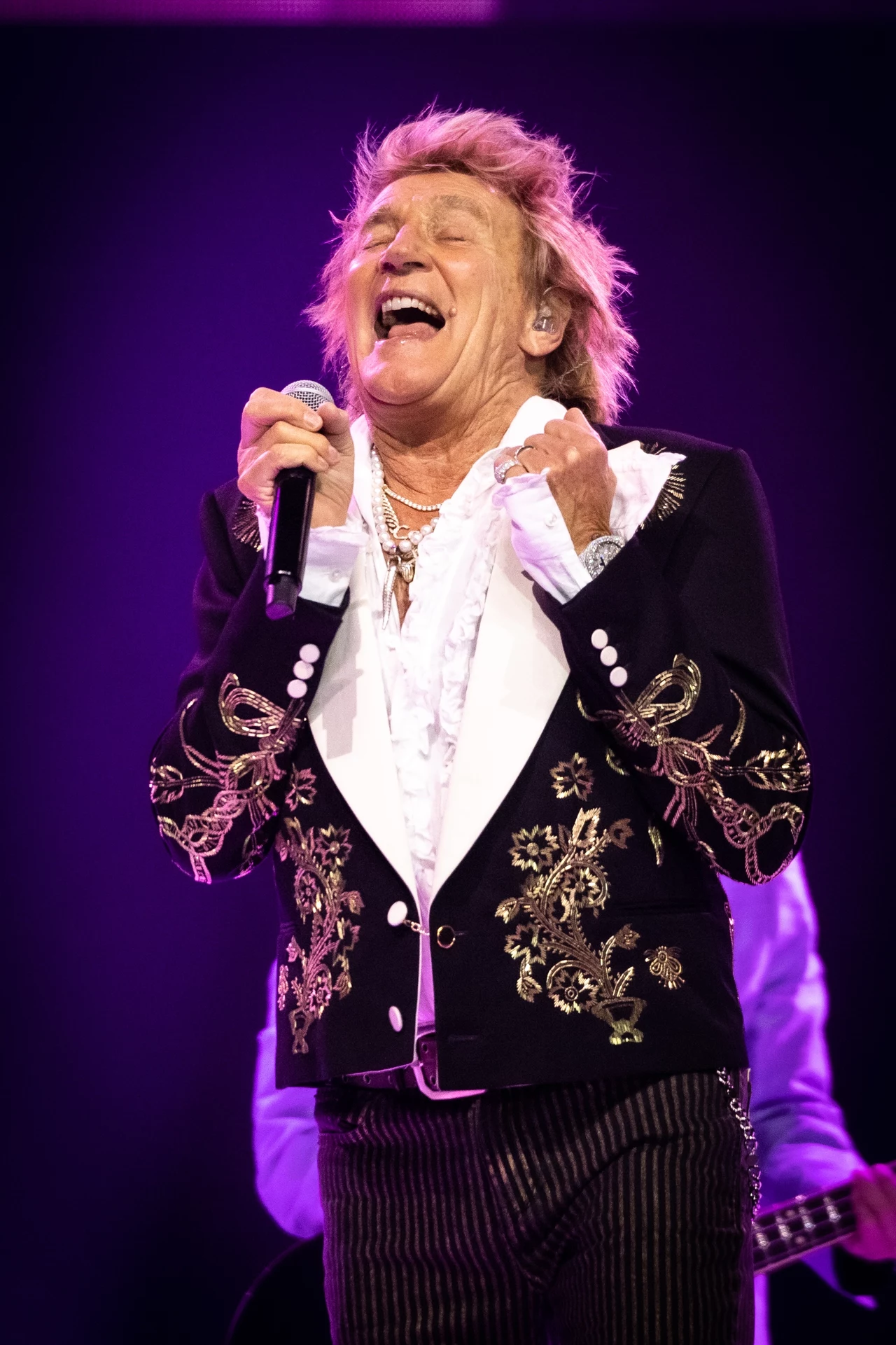 Rod Stewart 10 stycznia skończył 80 lat Osoba na scenie, trzyma mikrofon, ubrana w elegancką kurtkę z haftami, intensywnie śpiewa. Oświetlenie w tle jest fioletowe, co dodaje koncertowi dynamicznego charakteru.