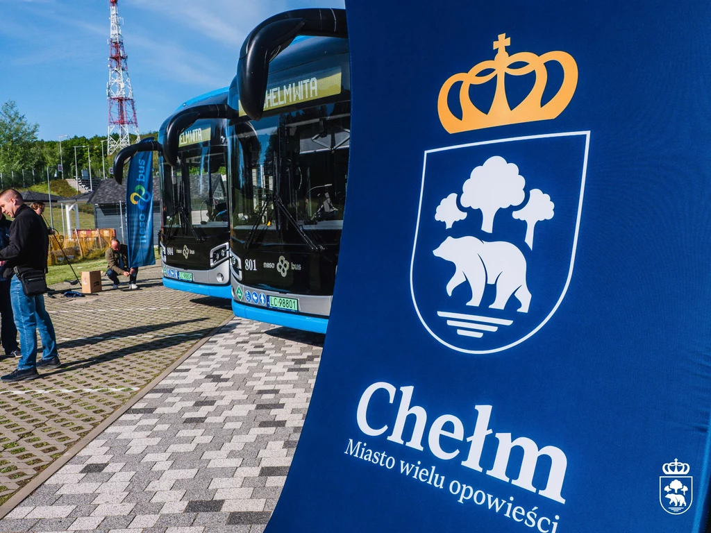 Chełm zamówił łącznie 26 NesoBusów Autobusy miejskie na przystanku obok tablicy z herbem miasta Chełm w słoneczny dzień. Na tablicy znajduje się wizerunek niedźwiedzia i korony oraz napis Chełm Miasto wielu opowieści.