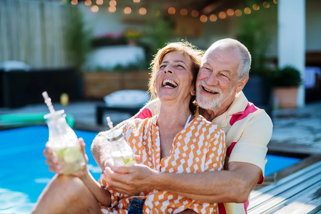 Wyjedźcie razem w piękne, nowe miejsce, zabierzcie znajomych i wspólnie spędźcie wspaniały weekend Para seniorów uśmiecha się i siedzi przy basenie, trzymając szklane butelki z napojem. W tle widać ogród z roślinami oraz drewnianymi elementami dekoracyjnymi. Oboje wyglądają na radosnych i zrelaksowanych.