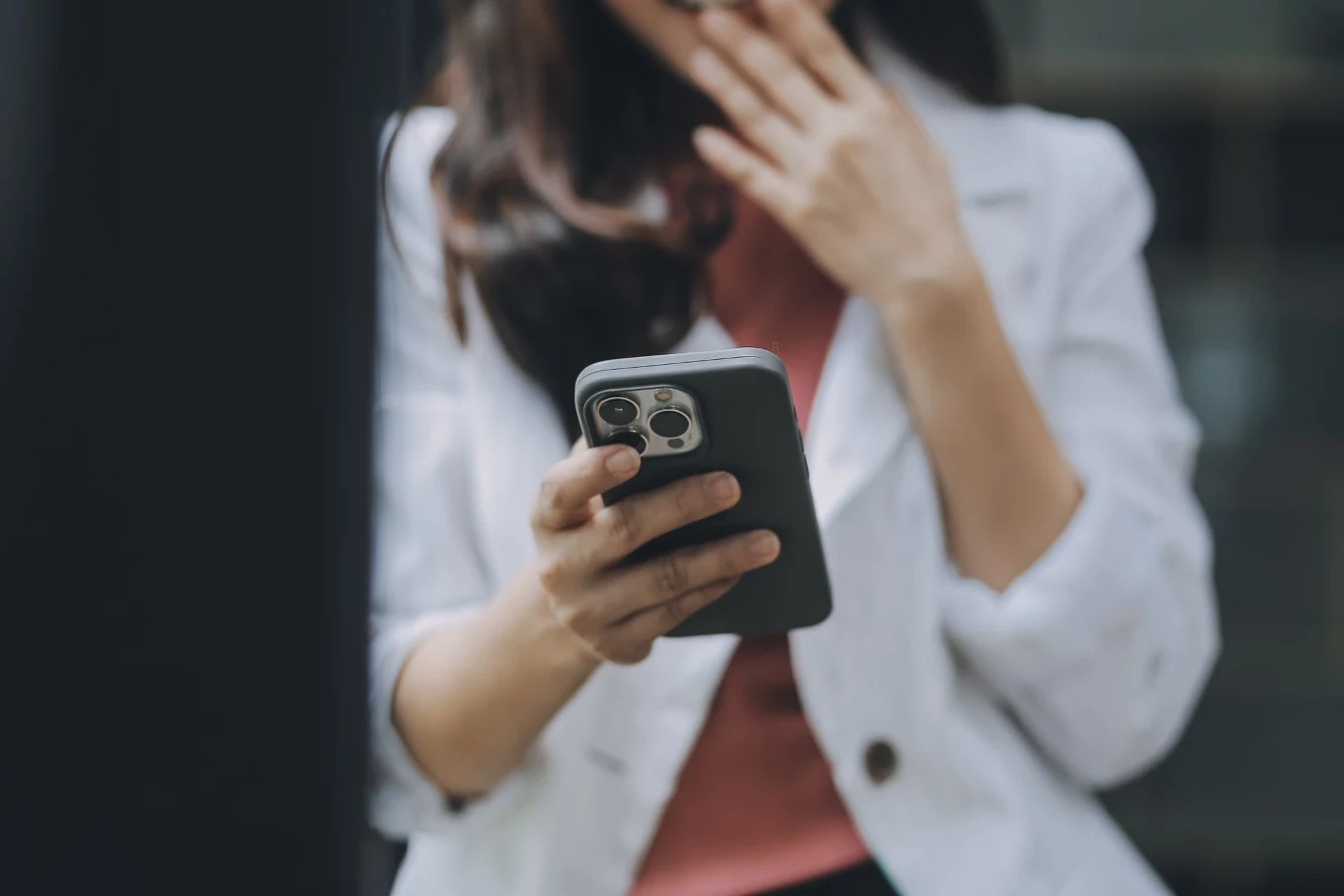 W pierwszej kolejności aktualizacja trafi do użytkowników urządzeń Apple. Osoba trzymająca smartfon w dłoni, lekko zakrywając usta dłonią, ubrana w jasną marynarkę. Widoczne są detale urządzenia i wyraz twarzy sugerujący zaskoczenie lub rozbawienie.