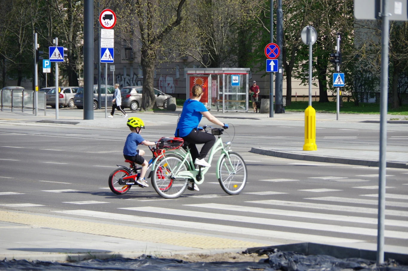 Kiedy rowerzysta ma pierwszeństwo? Kobieta i dziecko jadą na rowerach przez przejście dla pieszych na miejskiej ulicy, w tle widoczna budka telefoniczna i elementy infrastruktury miejskiej.