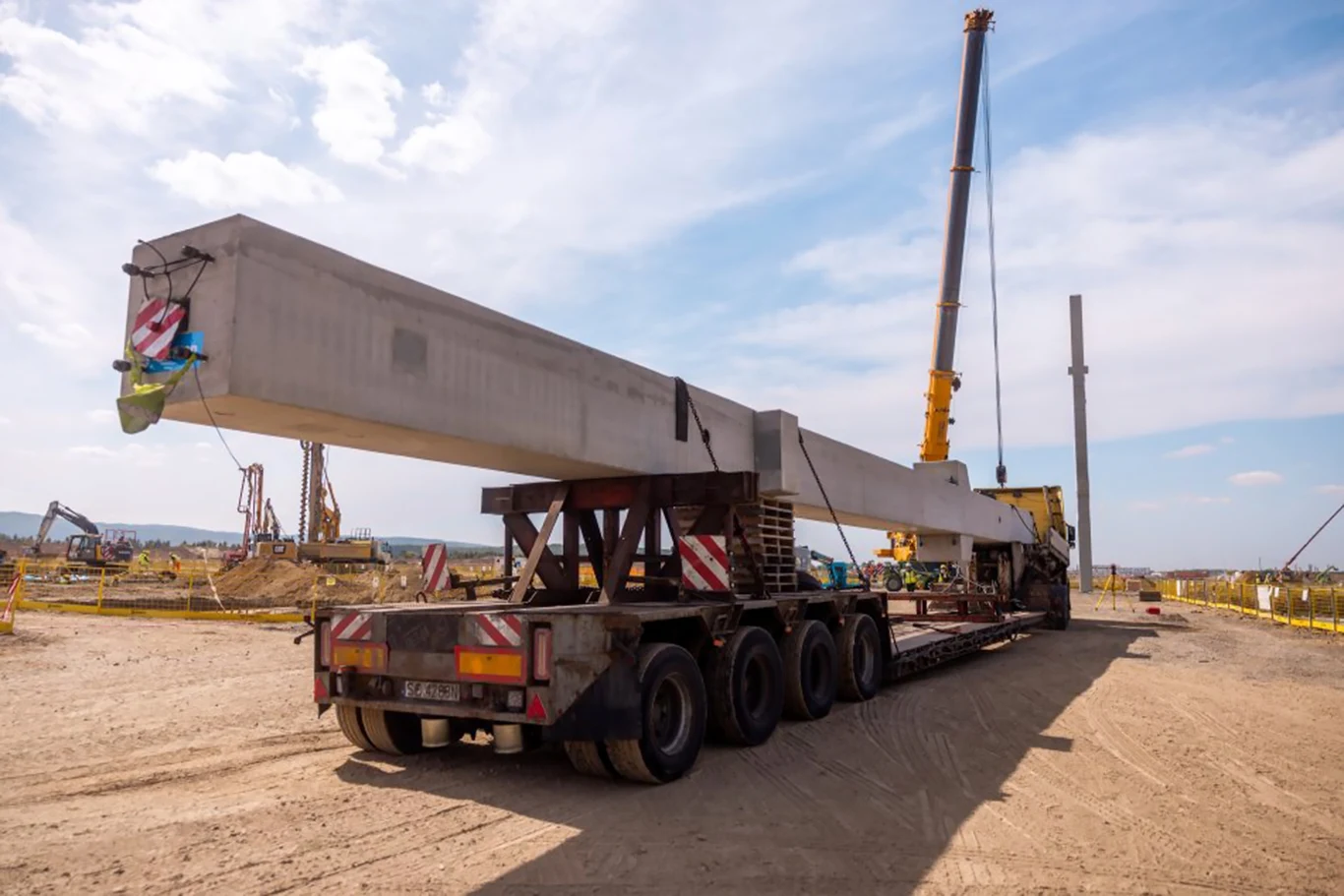 Transport długiej betonowej belki za pomocą specjalistycznej naczepy ciężarowej na placu budowy z dźwigiem w tle.