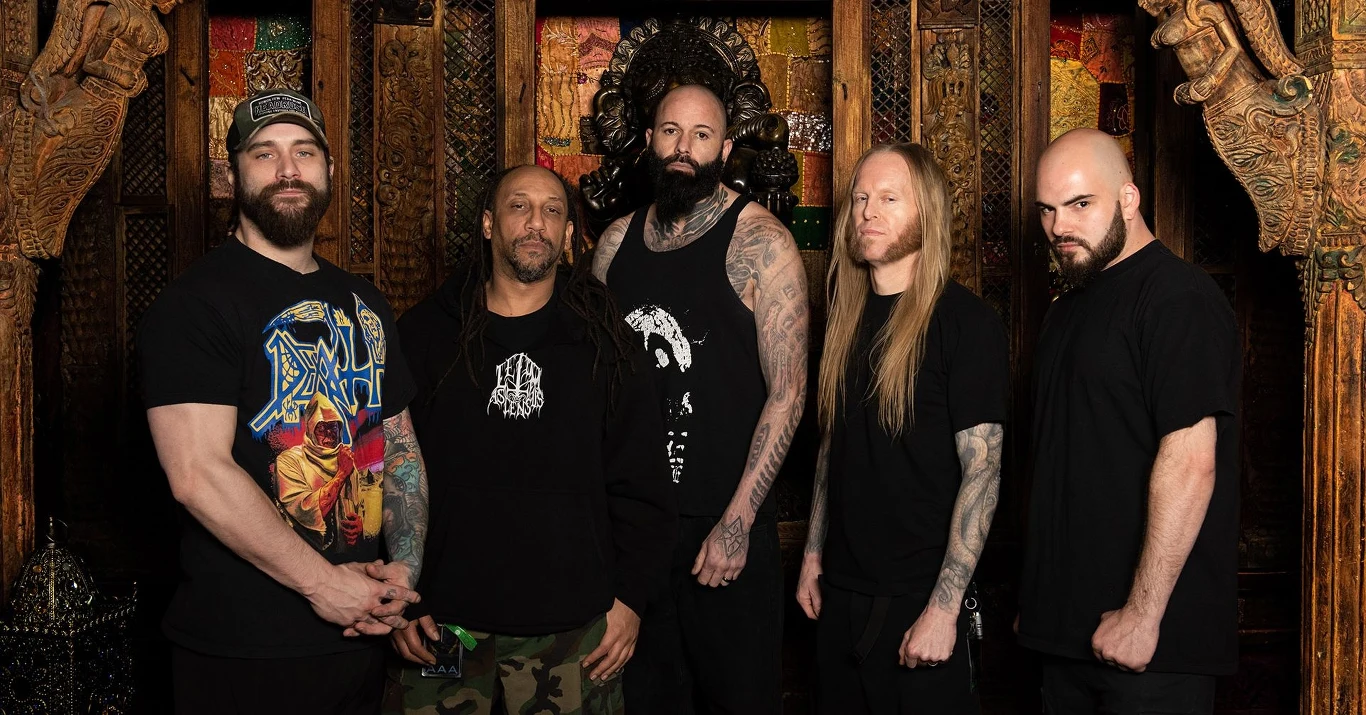 Suffocation powraca do Polski Pięciu mężczyzn stoi na tle ozdobnej, drewnianej ściany z motywami orientalnymi. Każdy z nich ubrany jest w ciemne stroje, a niektórzy mają tatuaże na rękach.