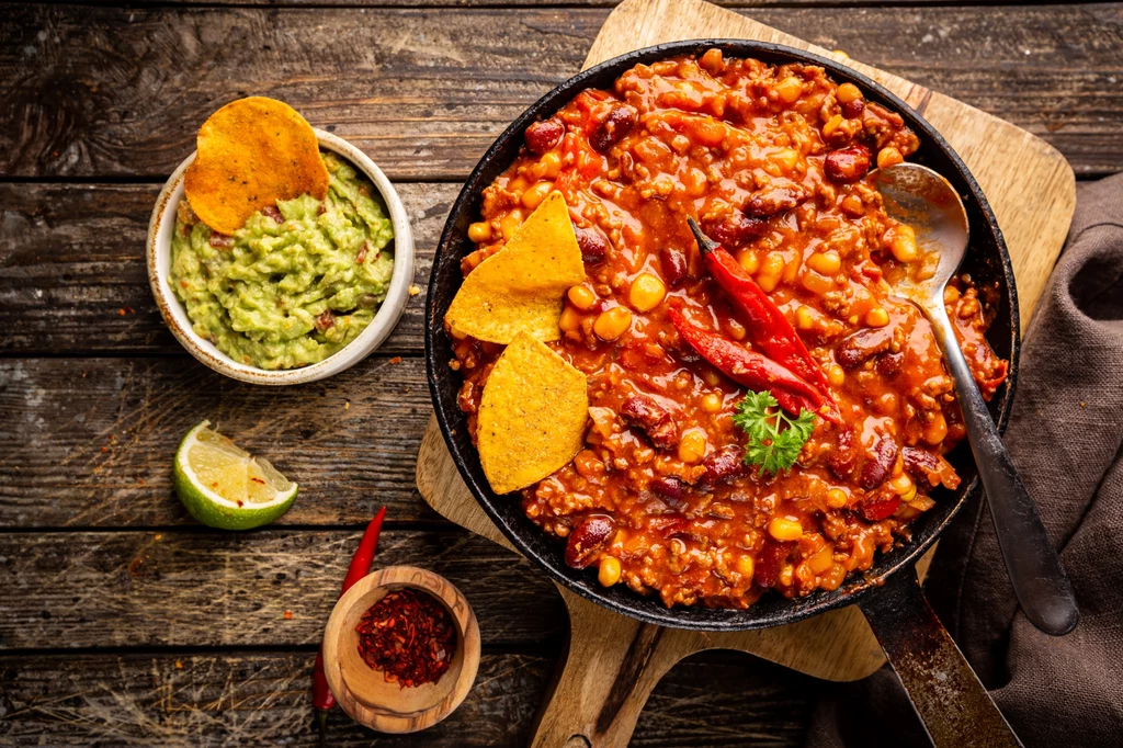 Może na jutrzejszy obiad przygotujesz chili con carne? Ta rozgrzewająca potrawa z pewnością przysłuży się twojemu zdrowiu Danie w stylu meksykańskim w żeliwnej patelni na drewnianej desce, z dodatkiem czerwonej fasoli, kukurydzy i mielonego mięsa, ozdobione papryczką chili i nachosami. Obok miseczka guacamole i plasterek limonki.