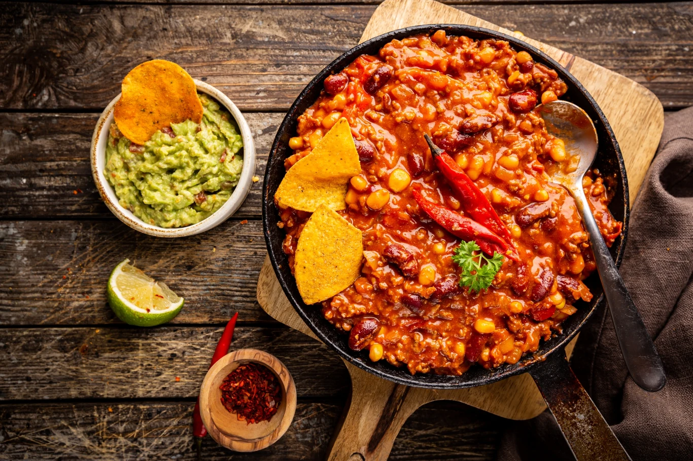 Danie w stylu meksykańskim w żeliwnej patelni na drewnianej desce, z dodatkiem czerwonej fasoli, kukurydzy i mielonego mięsa, ozdobione papryczką chili i nachosami. Obok miseczka guacamole i plasterek limonki.