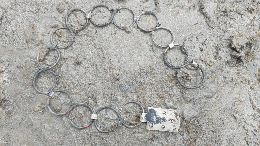 Naszyjnik odkryty w kraterze Metalowe kółka połączone w okrąg, z zamkiem, spoczywające na błotnistej powierzchni.