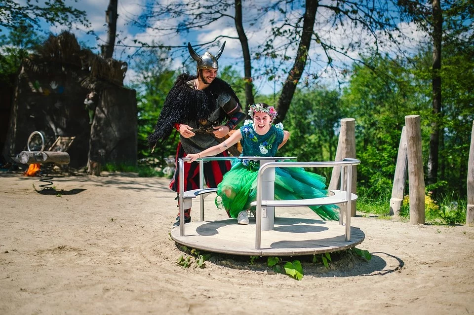 19 czerwca przygotujmy się na wspólny Dzień z Rusałkami Osoby przebrane w kostiumy Vikinga oraz wróżki bawią się na karuzeli na świeżym powietrzu w parku. W tle widać naturalną scenerię z drzewami i prowizoryczną drewnianą strukturą. Atmosfera jest radosna i beztroska.