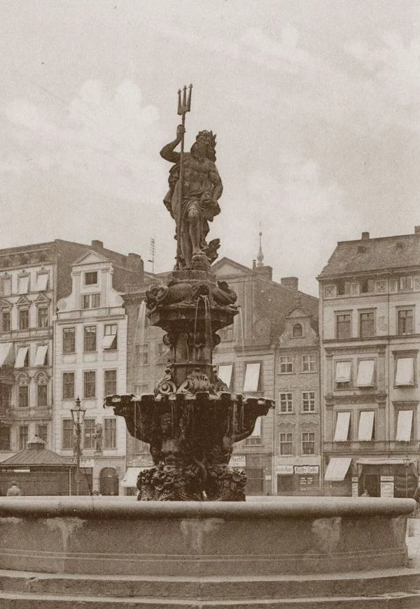 Fontanna Neptuna na placu, dawny symbol Wrocławia Fontanna z rzeźbą Neptuna na tle zabytkowych kamienic miejskich. Rzeźba przedstawia Neptuna z trójzębem, stojącego na ozdobnym cokole, otoczonego detalami architektonicznymi starego miasta. W tle widoczne są okiennice i dachy budynków.