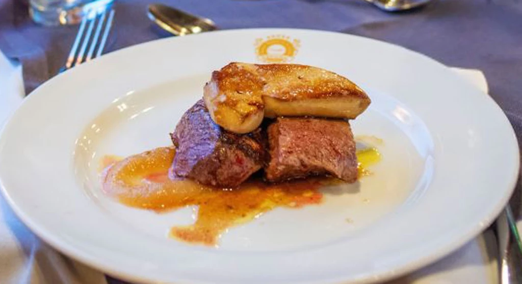 Polędwica wołowa z wątróbką "foie gras" serwowana w wagonie restauracyjnym luksusowego pociągu Transcantábrico Gran Lujo, Hiszpania. Danie składające się z kawałka soczystej, dobrze przyrządzonej wołowiny, ozdobionej kawałkiem foie gras, podane na eleganckim talerzu. Na stole widoczne są sztućce, sugerujące gotowość do spożycia tego wykwintnego posiłku.