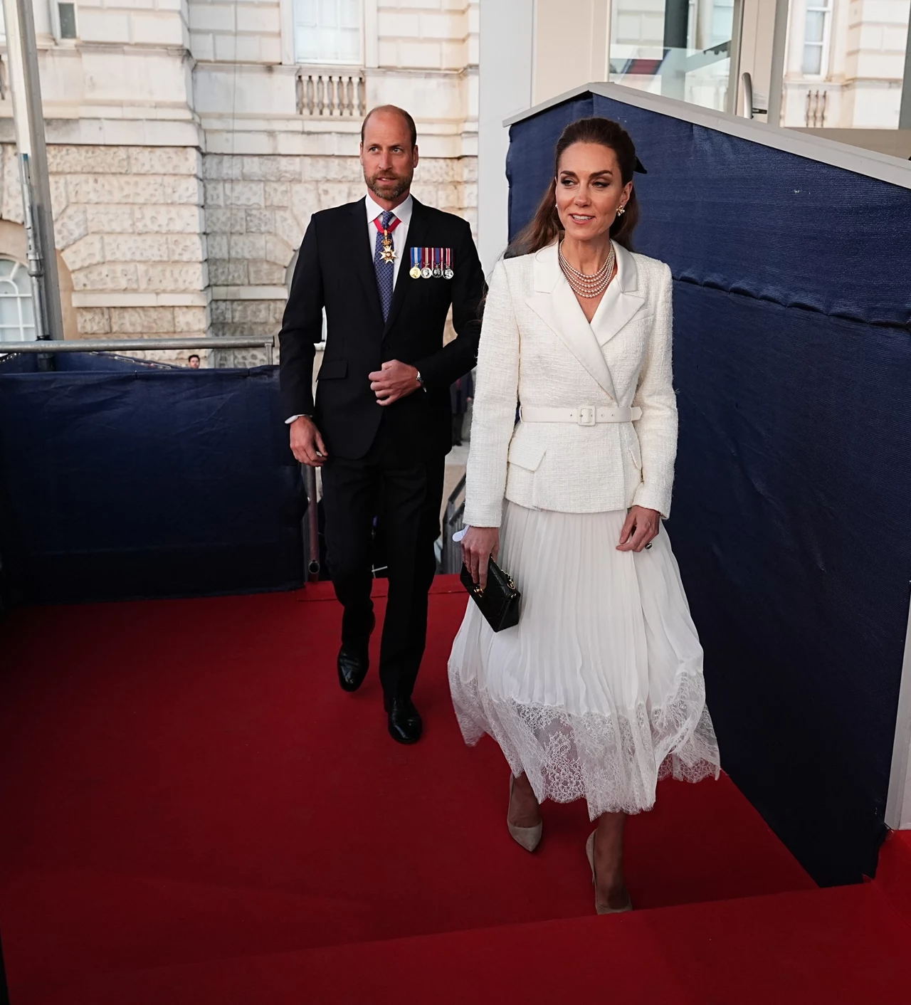 Na koncercie w Horse Guards Parade księżna także zachwyciła wyborem swojej stylizacji Elegancko ubrana kobieta w białej sukni i mężczyzna w garniturze z medalami spacerują po czerwonym dywanie. W tle widać fragmenty budynku i ogrodzenia zabezpieczającego.