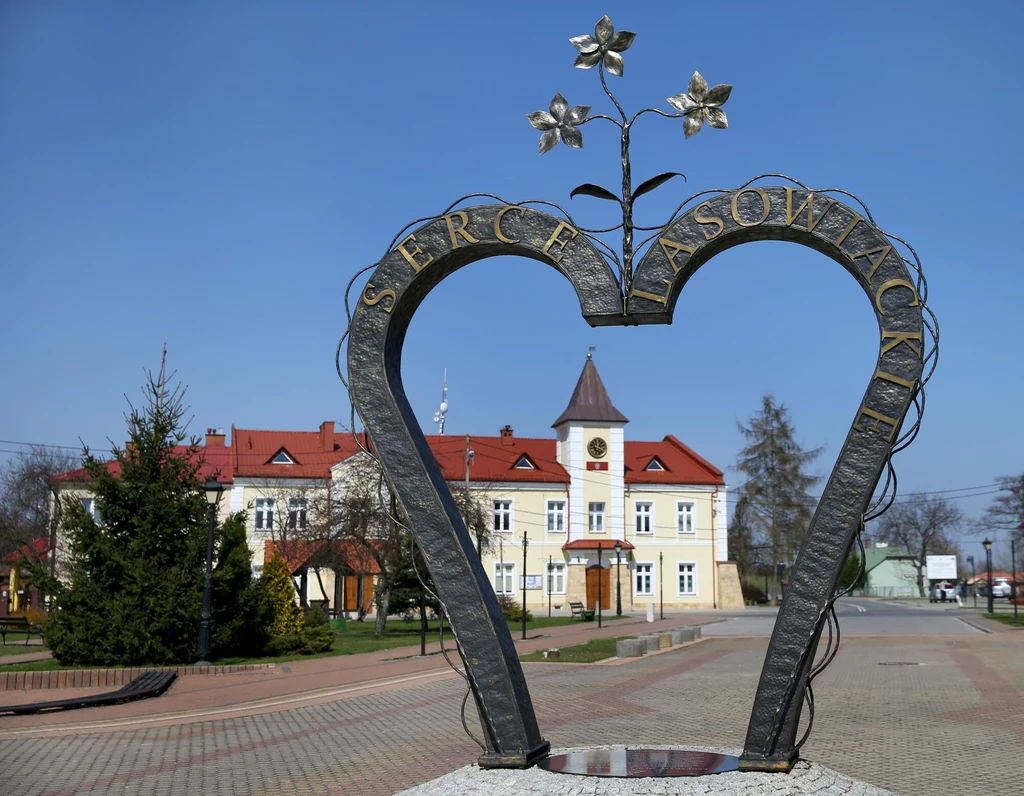 Rzut oka na Baranów Sandomierski Metalowa konstrukcja w kształcie serca z napisem 'Serce Lasowiackie', w tle XIX-wieczny budynek z czerwoną dachówką i wieżą zegarową, otoczony drzewami. Słoneczny dzień, błękitne niebo.