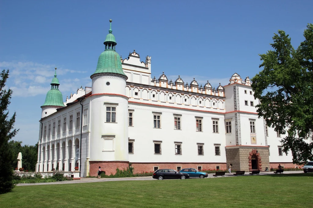 Prace nad budową zamku rozpoczęły się w 1591 roku Zamek z białymi murami i zielonymi dachami wież, otoczony zadbanym trawnikiem i drzewami, na tle jasnego nieba.