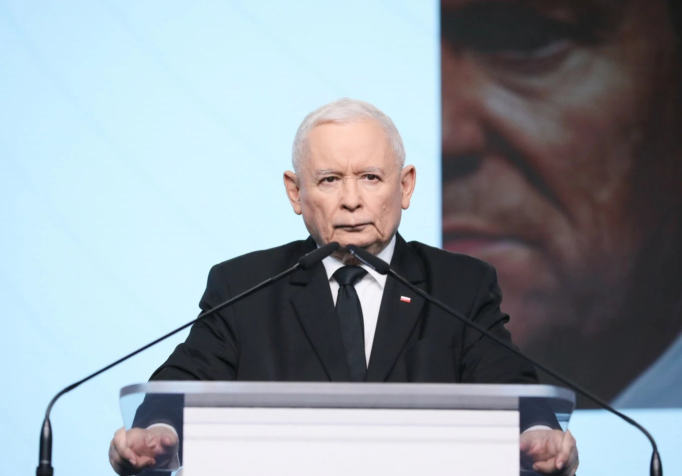 Jarosław Kaczyński, a w tle zdjęcie Donalda Tuska. To już ostatnie takie starcie? Mężczyzna w garniturze stoi przy mównicy, przemawiając do zgromadzonych. W tle widoczny jest duży ekran z fragmentem portretu.
