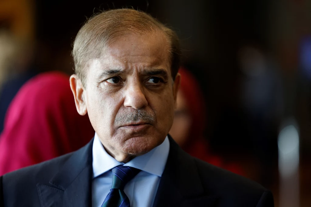 Premier Pakistanu Shehbaz Sharif Mężczyzna w średnim wieku w eleganckim garniturze i krawacie, z poważnym wyrazem twarzy, stoi w pomieszczeniu z rozmytym tłumem w tle.