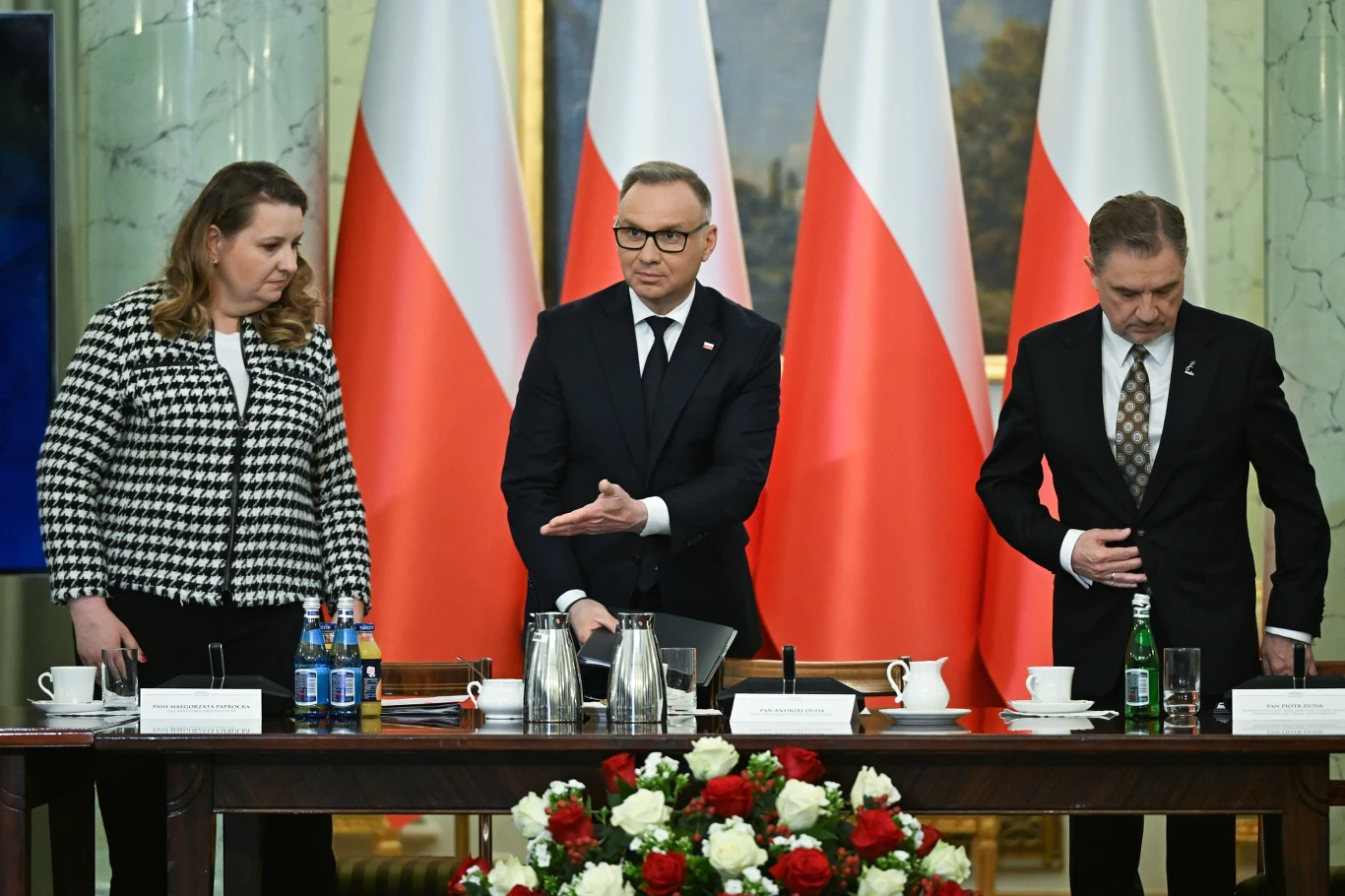 Andrzej Duda zawetował ustawę o obniżce składki zdrowotnej dla przedsiębiorców Trzy osoby za stołem w sali konferencyjnej z polskimi flagami w tle. Mężczyzna w środku gestykuluje, wskazując na coś na stole. Przed nimi duży stół z kwiatami i butelkami wody mineralnej.