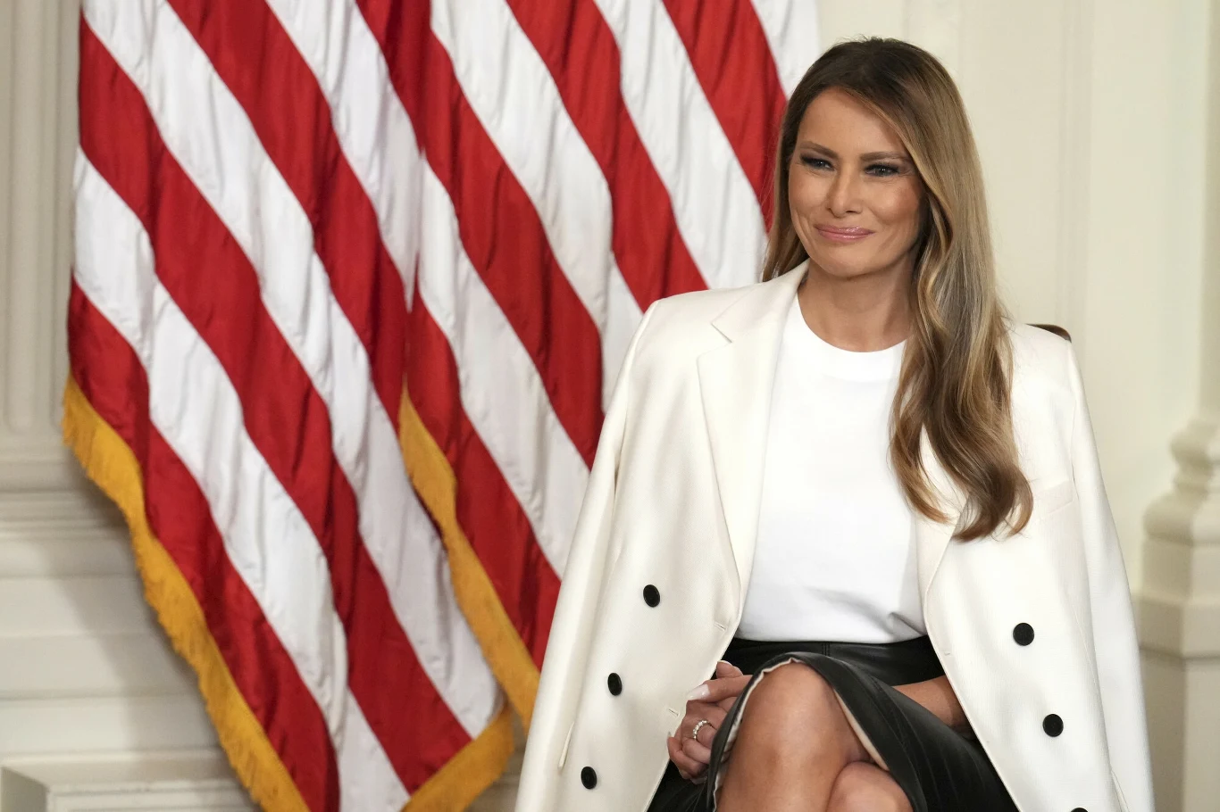 Melania Trump została gospodynią jednego z wydarzeń w Białym Domu Kobieta siedzi przed flagą USA, ubrana w elegancką białą marynarkę oraz czarno-białą spódnicę. Ma długie, jasnobrązowe włosy i lekki uśmiech na twarzy.