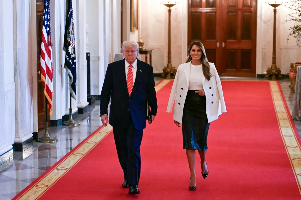 Stylizacja Melanii Trump to urzeczywistnienie klasyki i elegancji Mężczyzna i kobieta idą eleganckim korytarzem z czerwonym dywanem, obok flag USA przy białej ścianie. Mężczyzna ma ciemny garnitur i krawat, kobieta nosi białą marynarkę i czarną spódnicę.