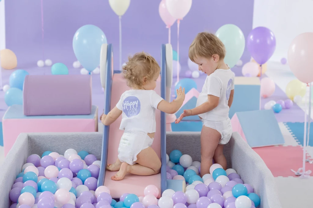 Pieluszka Bella Happy Soft&Delicate z kanalikami sprawi, że nasza podróż będzie spokojniejsza Dwoje małych dzieci bawi się w basenie z kulkami w pastelowych kolorach. Dzieci noszą białe ubranka, a wokół nich znajdują się różnokolorowe balony oraz miękkie elementy do zabawy. Cała scena ma przyjazny i beztroski charakter.