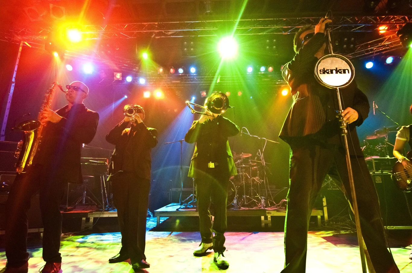 Skankan zagra koncert w Krakowie Grupa muzyków na scenie, grających na instrumentach dętych, oświetlonych kolorowymi reflektorami. Atmosfera koncertowa z intensywnym oświetleniem i żywymi kolorami tworzy dynamiczny nastrój.