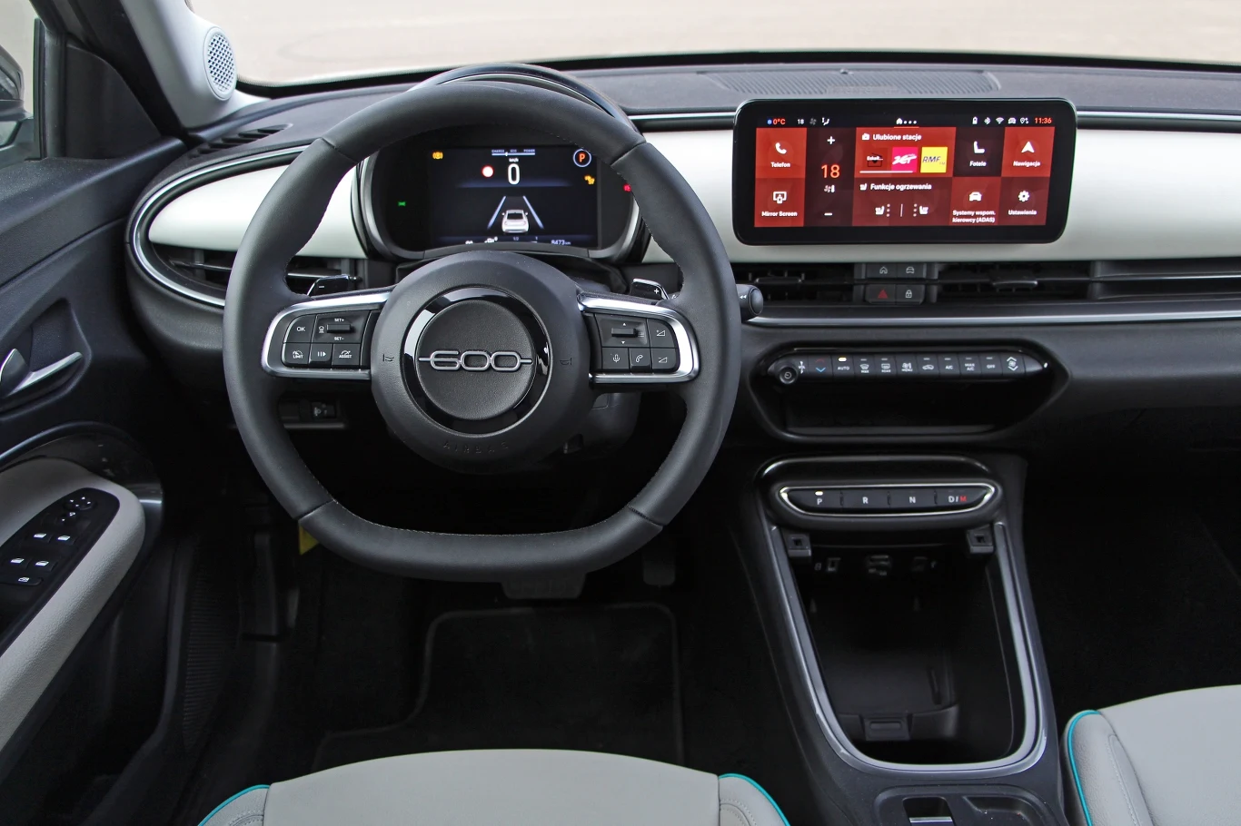 Wnętrze samochodu elektrycznego z centralnym wyświetlaczem na desce rozdzielczej oraz systemem multimedialnym i kierownicą z logo marki. Widoczne są przyciski sterowania na kierownicy i panelu bocznym.