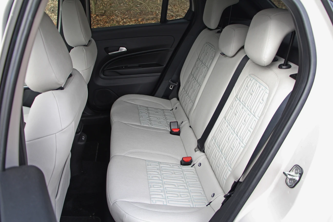 Widok wnętrza samochodu z białymi, skórzanymi siedzeniami, obejmujący tylną kanapę z pasami bezpieczeństwa i jasnym wykończeniem. Przyciemnione szyby oraz elementy drzwi dodają elegancji i nowoczesności.